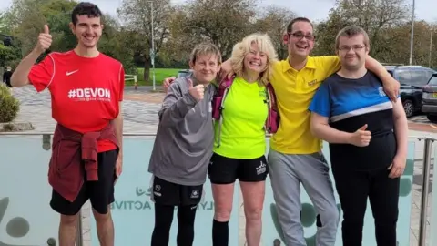 Ali Cattle A group of five people smiling with their arms around each other's shoulders giving thumbs up signs while wearing running clothing and trainers.