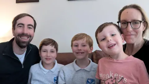 Calum Dad Calum, his sons Archie, Andrew, Roddy and mum Katie are sat side by side on a couch smiling at the camera. Archie and Andrew are in their school uniform and Roddy is wearing a top with monkeys on it.