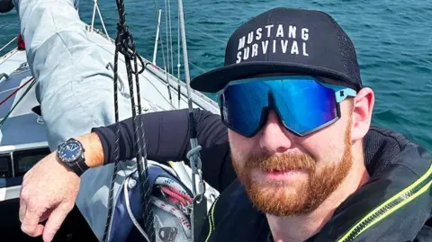 Kyle Meredith Kyle is on a boat and wears a navy blue long-sleeved top, blue sunglasses and a navy blue cap