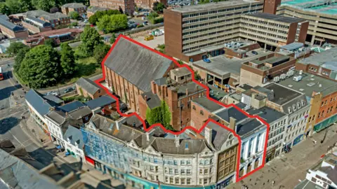 West Northamptonshire Council Three-storey buildings in a town centre at a curved intersection between two streets. The Corn Exchange site has a white entrance, and a large brick hall at the rear.