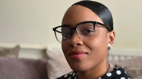 BBC Dainya in a black and white polka dot blouse. She wears a necklace, earrings and glasses. She is sat on a sofa looking at the camera.