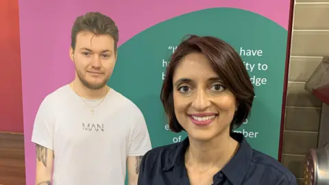 A woman stands in front of a poster inside a building. The woman has short dark brown hair and she is wearing a dark blue shirt. The pink and green poster has a photo of a man wearing a white T-shirt on it.