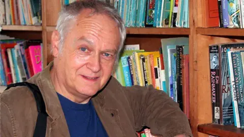 BBC Phil Rickman, who has short grey hair and blue eyes, standing next to a bookcase. He's wearing a brown jacket and blue jumper