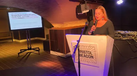 BBC Carly Heath holding a microphone and standing at a white lectern that says Bristol Beacon on it. There is a big screen in the background giving info about the Bristol Music Fund