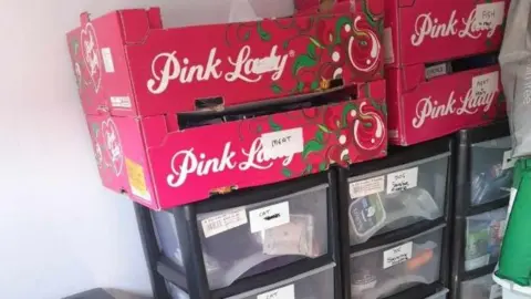 Wendy Isaacs Containers and cardboard boxes in Wendy Isaacs' spare room, where she stores supplies for the pet food bank. Four Pink Lady apple boxes are stacked near the wall, labelled with their contents. Drawers can be seen below them, also labelled with the contents.
