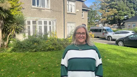 Cheryl wears a green and white striped jumper standing on the grass in front of a grey stone house. She has blonde long hair and is wearing black framed glasses and a gold chain.