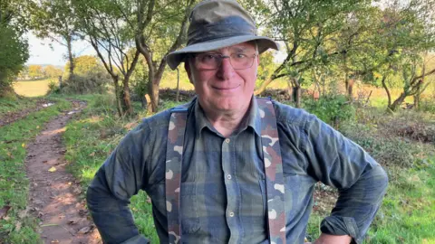 BBC Werner Lothmann, who is wearing a green hat, green shirt and camouflage braces, standing in a green space at Langdon Hospital, Dawlish.