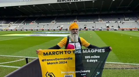 Manny Singh Kang Manny Singh Kang with banner saying "March to the Magpies" on a football pitch inside a stadium. He wears a yellow head covering and has a long white beard