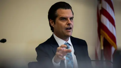 Getty Images Matt Gaetz, wearing a suit, gestures with his first hand while sitting and speaking at a microphone during a hearing