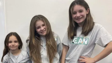 BBC The three sisters are looking to camera in grey tops and smiling.