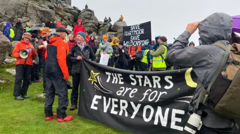 BBC A large group of people wearing camping gear stand in front of Hound Tor. Some are holding a large banner that says 'The stars are for everyone'. In the background another banner says 'Save Dartmoor, save wild camping'.
