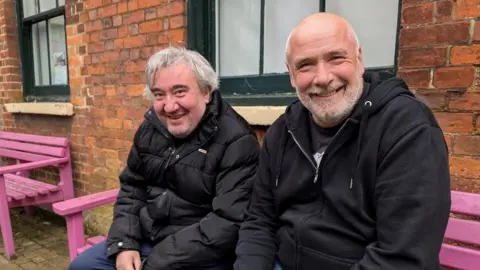 Tara Dolby/BBC Two men, Lee and Steve, are sitting on a bright pink bench smiling at the camera. Lee has grey stubble and grey hair, he is wearing a black jacket and blue trousers. Steve has a shaved head and white and grey cropped beard. He is wearing a black hoody over a black T-shirt.