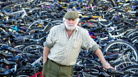 Graeme Hart/PPA Nigel Carter leans against one bike as he stands amongst thousand while looking at the camera.
He has white hair and a beard, wearing a checked shirt and green trousers.