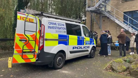 The back of a police Crime Scene Investigation van with one of its rear doors open and the word "police" in blue lettering down the side of a white panel. Pupils and a teacher stand in the background.