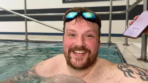 BBC Jordan Brooks smiles in the swimming pool, his goggles on his head. He has brown hair and a beard, with tattoos on his upper arms.