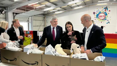 BBC Ex-PM Gordon Brown with The Junction's Beth Major and John Boumphrey from Amazon at the new Multibank centre in Teesside. They are looking at cardboard boxes filled with