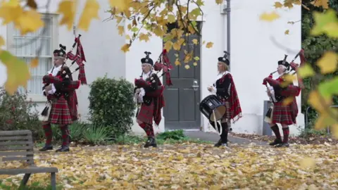 Sheila Hatcher and Denise Morrison stood outside in their uniforms, consisting of a black velvet jacket, red tartan bagpipes, a black drum, white lace collars and black hats