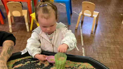 Home-Start Wellingborough & District A baby girl wearing a grey top pours orange and green coloured rice granules into a green cup