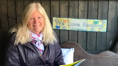 Paula Flood Paula Flood, wearing a brown leather jacket and a white and pink scarf, sits on a wooden bench. She is smiling at the camera. A sign behind her says "We love reading!"