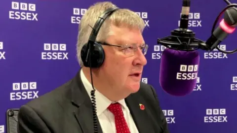 Roger Hirst with grey hair wearing a black suit jacket, a white shirt, and a red tie.