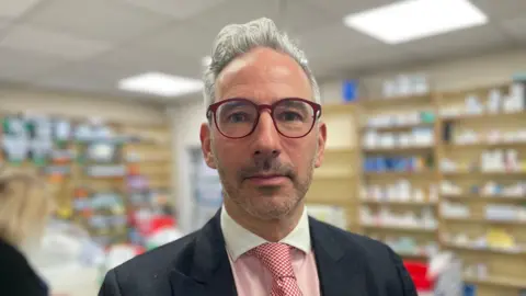 BBC/Gemma Dillon Ashley Cohen, wearing glasses, stands in a pharmacy wearing a white-collared shirt with red tie.
