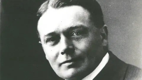 Bournville Village Trust A black and white photo of a man in a suit and shirt. He has short dark hair, swept back and centre-parted and is clean shaven.