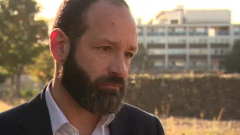 BBC Southwark Council leader Kieron Williams in a striped shirt and dark suit jacket. He has short dark hair, a moustache and beard. He stands in front of an out of focus building.