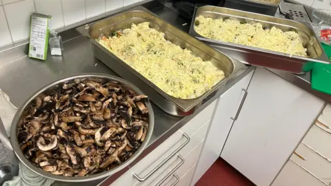 A large silver bowl with sliced mushrooms and a large rectangular serving dish with cheese and creamy food inside. The containers are on a stainless steel work surface in the community centre kitchen.