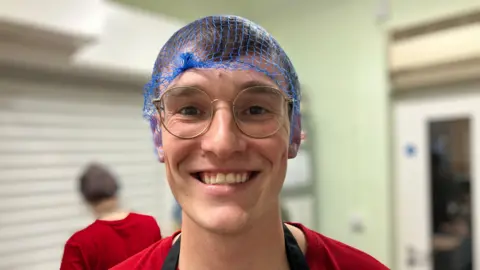 BBC A smiling man with round-framed glasses is wearing a blue hair net, red T-shirt and a black apron on top.