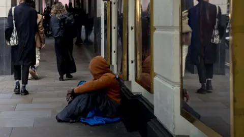 Getty Images Homeless person seen sitting on the ground along a London street. He is wearing a yellow hoodie and looking away from the camera keeping his face concealed. Pedestrians can be seen walking along the street.