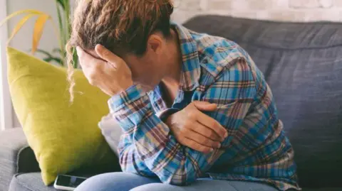Getty Images A woman in a blue shirt is sat on a sofa with her head in her hand. She looks upset, although you can not see her face. There is a yellow cushion next to her and a plant.