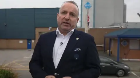 BBC Dave Fishwick standing outside a factory with blue cladding and brinde brick walls. He is wearing a white shirt and dark blue suit.