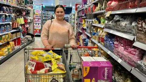 Tyesha Muncaster Tyesha Muncaster stands in the aisle of a supermarket. In front of her are two trolleys which are filled with pet food.