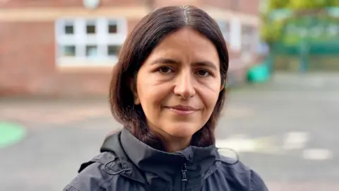 BRANWEN JEFFREYS/BBC A picture of Shabnam looking and smiling at the camera on the school playground. Her dark her is tucked into a black raincoat.
