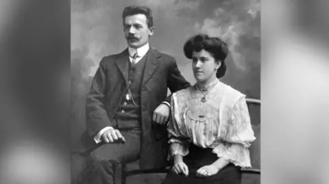Eddie Wulff Max and Mabel Wulff, a Welsh-German couple, are photographed in black and white. Sat side by side Max and Mabel are dressed smartly in a three piece suit and a white blouse with a skirt respectively.