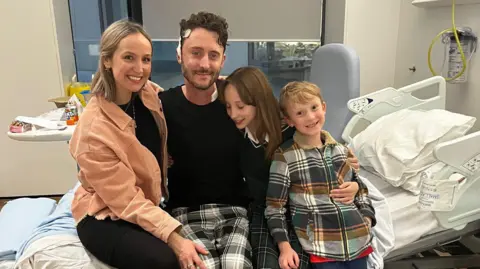 Matthew White Matthew White wearing pyjamas and sitting on a hospital bed, with his wife on his right hand side and his son and daughter on his left.
