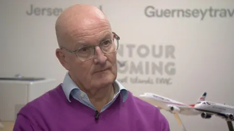 Robert Mackenzie is sitting in a room that has Jersey travel and Guernsey travel etched on the walls and there are two model aeroplanes to his left. He looks serious and is wearing a mic on his purple jumper.