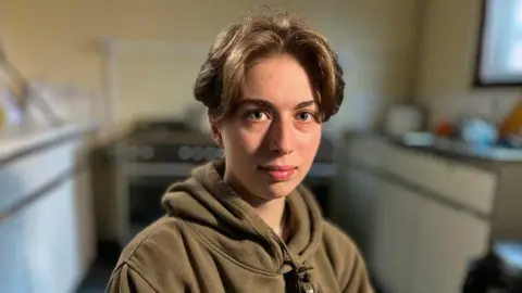 Martin Giles/BBC Aimee Clasby, trainee youth development worker, looks directly at the camera. She is wearing a brown hoodie.