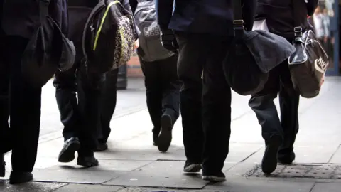 PA Media A group of school children walk down a street carrying their schoolbags. The photograph is taken from behind, as they walk away. They are all wearing dark clothing.