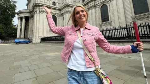 Canseecantsee Claire Sisk standing outside St Paul's Cathedral in London, she is standing on a pavement, with both her arms outstretched. She is holding a walking cane, with a red top. She is wearing jeans and a white T-shirt, a pink denim jacket and has a bag across her body. A blue car and St Paul's Cathedral can see been behind her.