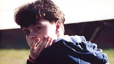 Family photo Picture of Emma-Jayne Knowles looking over her shoulder towards the camera with her hand in front of her face. She has brown wavy hair and is wearing a blue jacket.