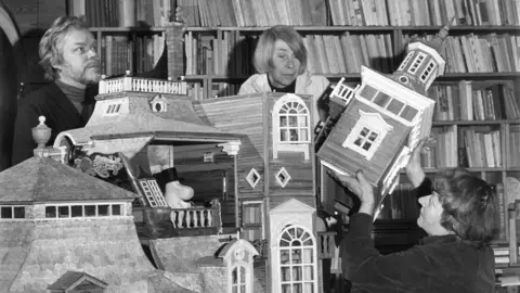 Per Olov Jansson Black and white image of three people standing in front of bookshelves, including Tove Jansson, as they construct a large dolls' house sized Moomin house