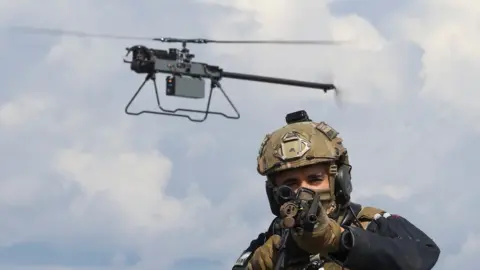 Royal Navy A Ghost drone hovers just behind an armed marine who is pointing his rifle at the camera