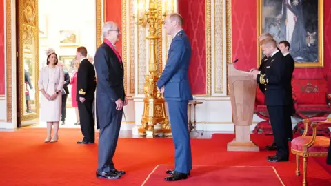 PA Media Sir Patrick speaking to Prince William as he receives his medal in an ornate red room in Buckingham Palace