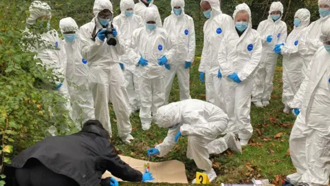BBC School children wearing white protective overalls and blue rubber gloves inspect a mock-up of a crime scene with the help of a police officer who is wearing a black coat and trousers.