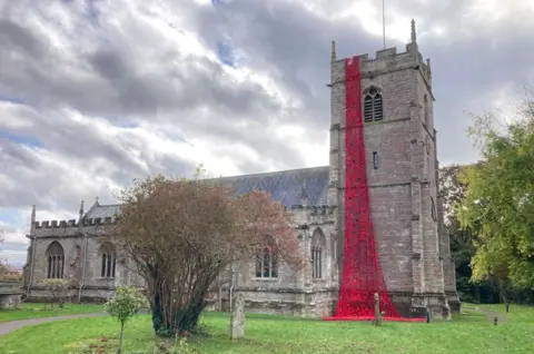 BBC A cascade of poppies on the church bell tower going from the top to the ground. The rest of the church is behind the tower and there are trees in the foreground and to the right.