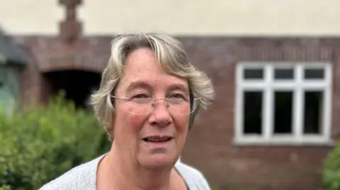 Christine Willoughby looking into the camera in front of a house. She has blonde grey hair, wears glasses and has a grey cardigan on.
