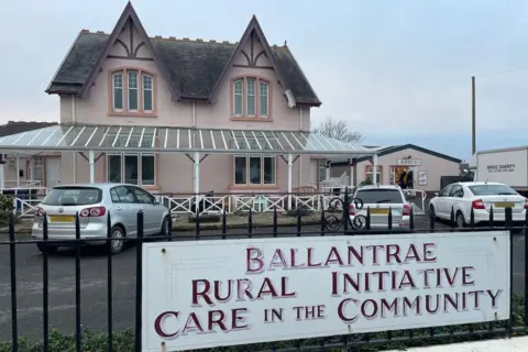 A large detached light pink house with a dark grey roof. Cars are parked in front of it and a black iron fence is in the foreground with a sign which says "Ballantrae Rural Initiative Care in the Community".