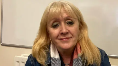 BBC A woman with long blonde hair wearing a red, white and black scarf and a denim jacket, sitting in front of a whiteboard.
