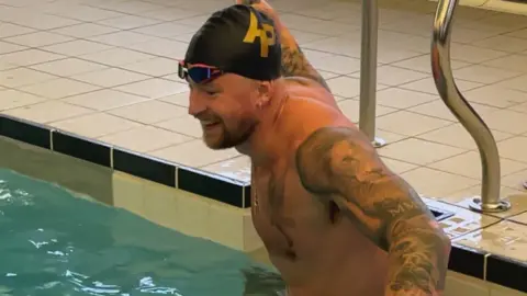 BBC Adam Peaty smiles as he climbs into a pool wearing a black swimming cap with AP in gold letters and swimming goggles on his forehead.